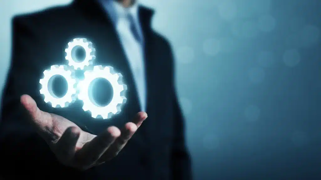 A businessman in a dark suit holding out his hand with three glowing white gears hovering above it, symbolizing digital automation, business process management, and system integration in a modern technology environment.
